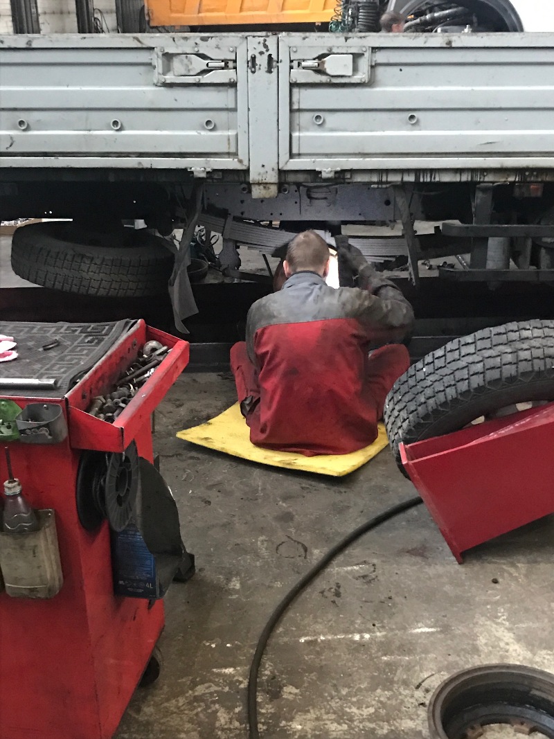 Рабочий ремонтирует грузовой автомобиль, сидя на коврике.